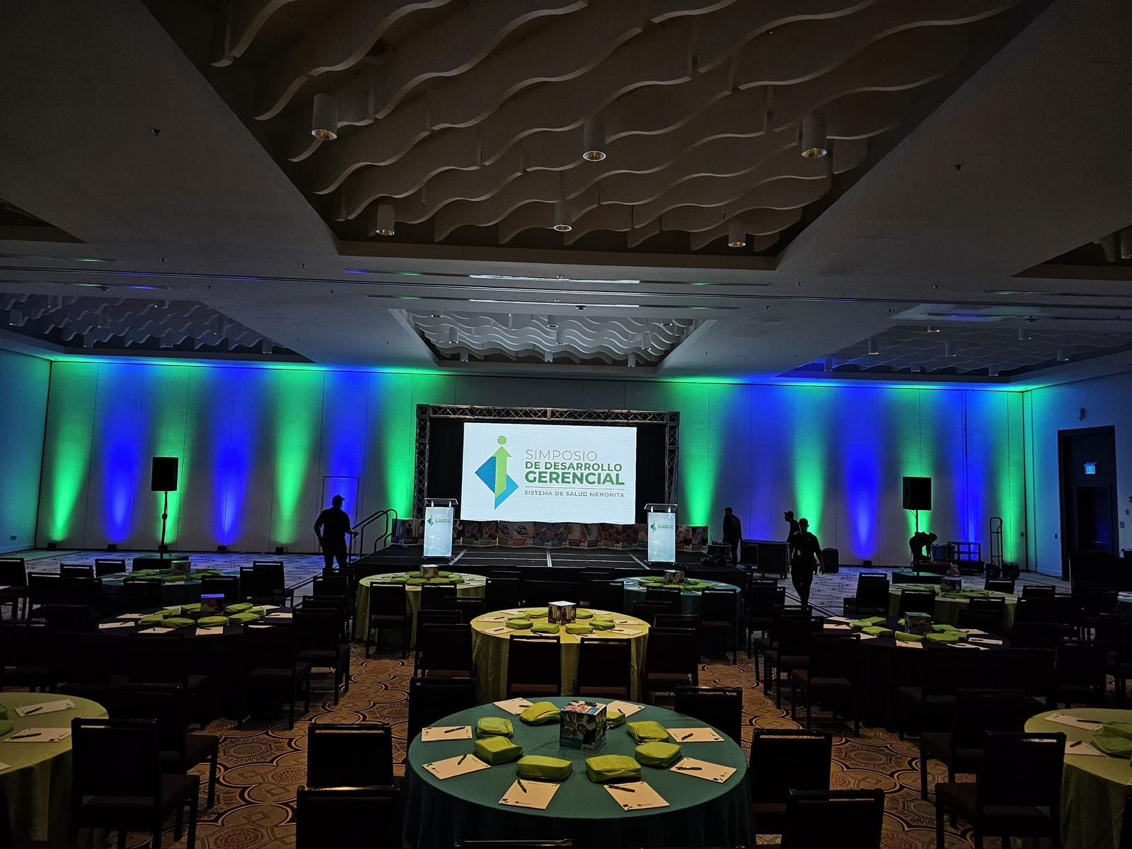 Green and blue uplighting frames a central screen and stage in a banquet room
