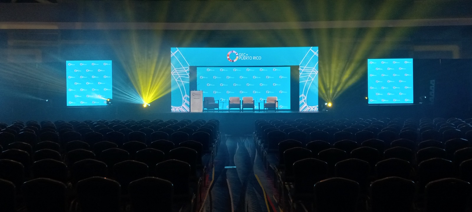 Three screens and yellow beams frame a conference stage